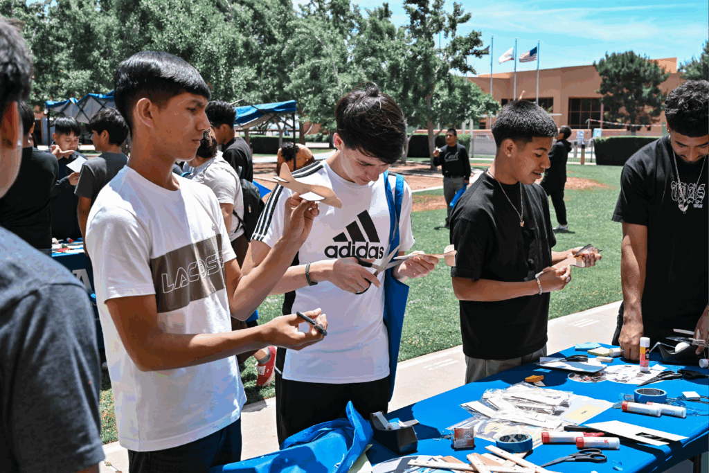 A group of students stands at an outdoor table, working with Balsa Glider STEM kit projects with various materials under a sunny sky.